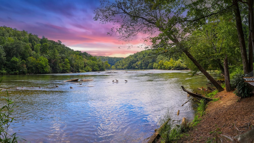 a panoramic of a gorgeous summer landscape on the Chattahoochee River with flowing water surrounded by lush green trees, grass and plants and powerful clouds at sunset in Atlanta Georgia USA