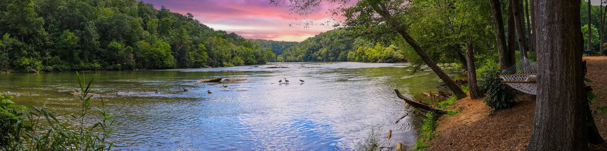 a panoramic of a gorgeous summer landscape on the Chattahoochee River with flowing water surrounded by lush green trees, grass and plants and powerful clouds at sunset in Atlanta Georgia USA