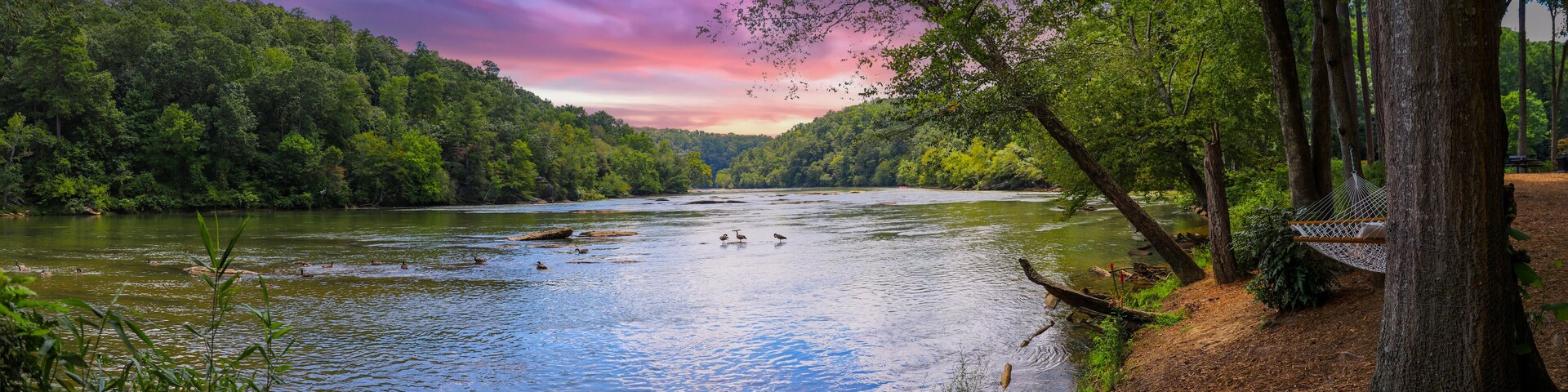 a panoramic of a gorgeous summer landscape on the Chattahoochee River with flowing water surrounded by lush green trees, grass and plants and powerful clouds at sunset in Atlanta Georgia USA
