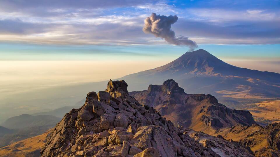 A mountain with a visible smoke plume, rocky terrain, and distant valleys.