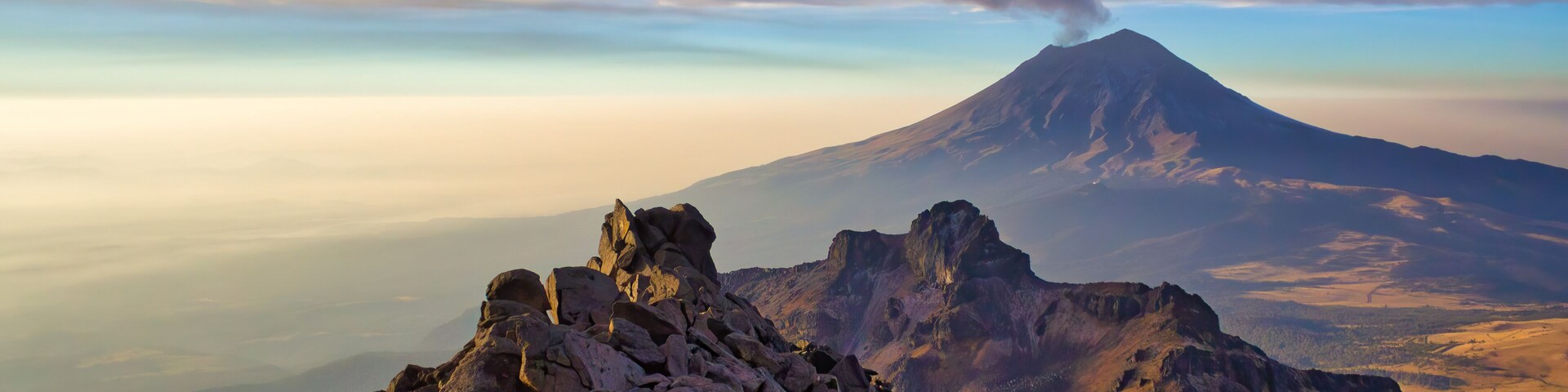 Una montaña con una columna de humo visible, terreno rocoso y valles lejanos.