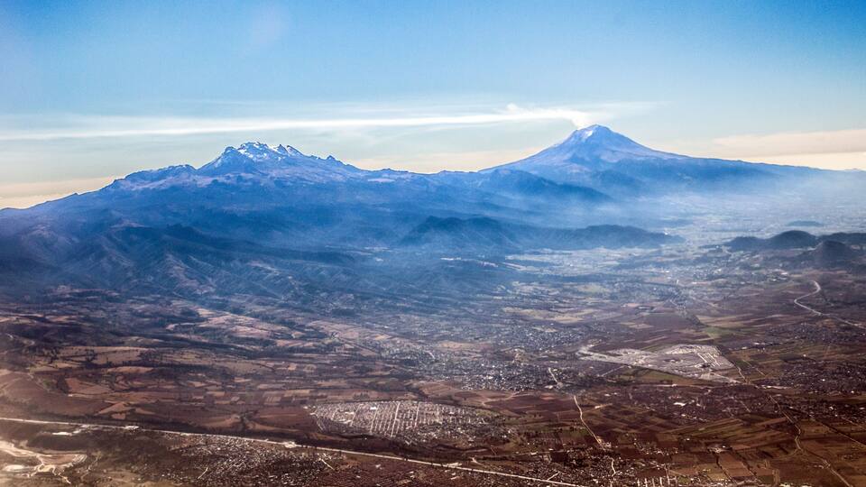 A mountain range with a prominent peak, a valley with agricultural fields, and a distant cityscape.