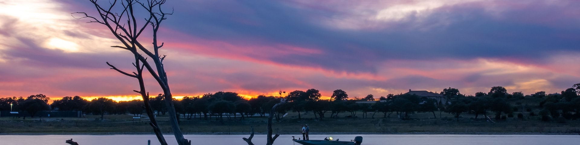 Un atardecer sobre un lago tranquilo con siluetas de árboles y una orilla distante.