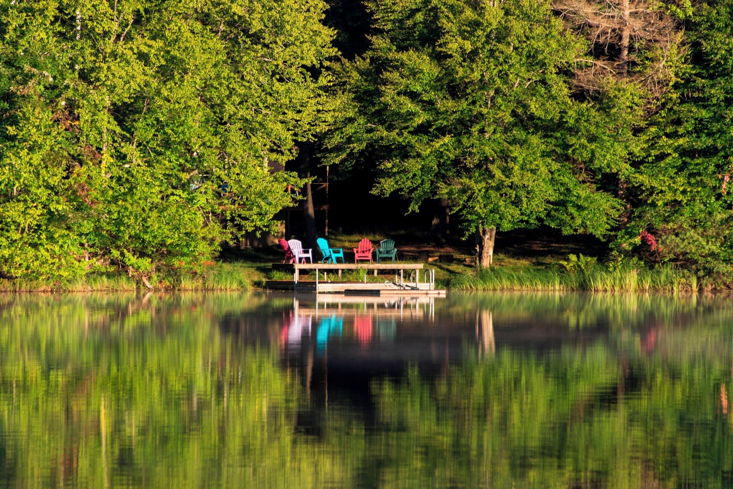 A calm lake with a dock and colorful chairs.
