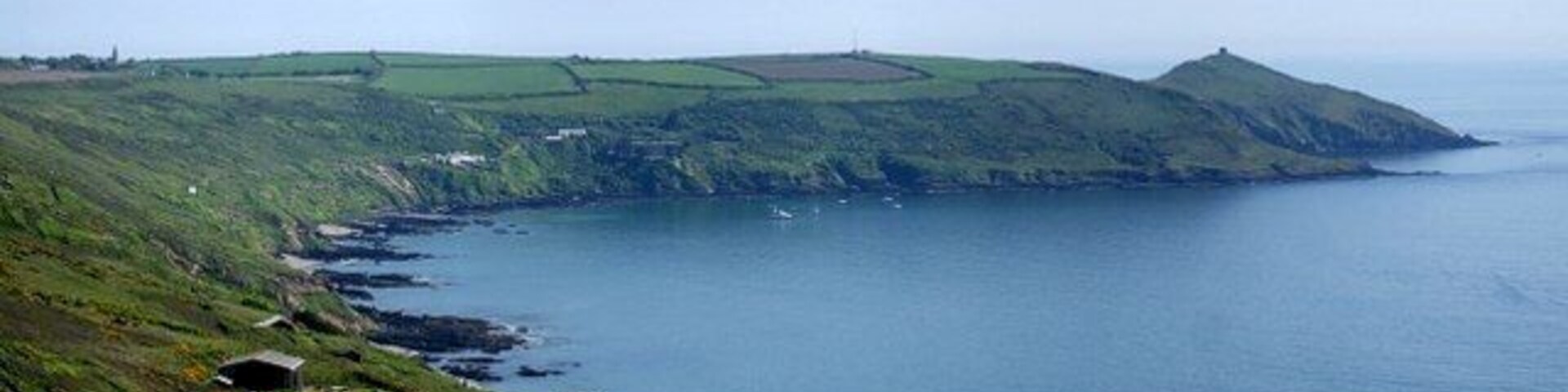 A coastal landscape with green hills, a rocky shoreline, and a small structure near the water.
