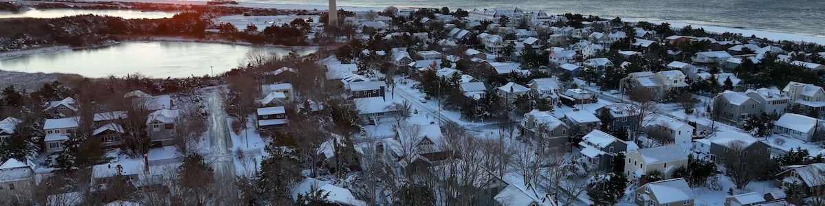 A coastal town at sunset with a lighthouse, snow-covered houses, and a frozen lake.