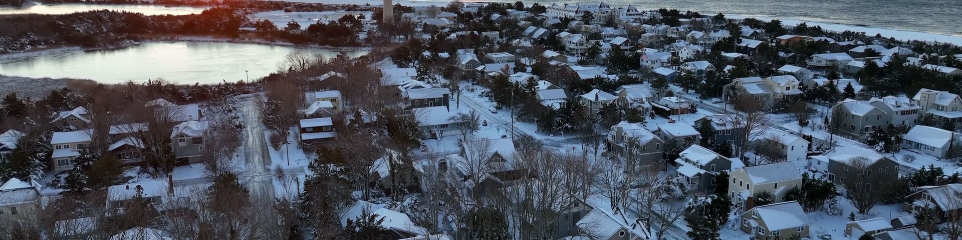 Un pueblo costero al atardecer con un faro, casas cubiertas de nieve y un lago congelado.