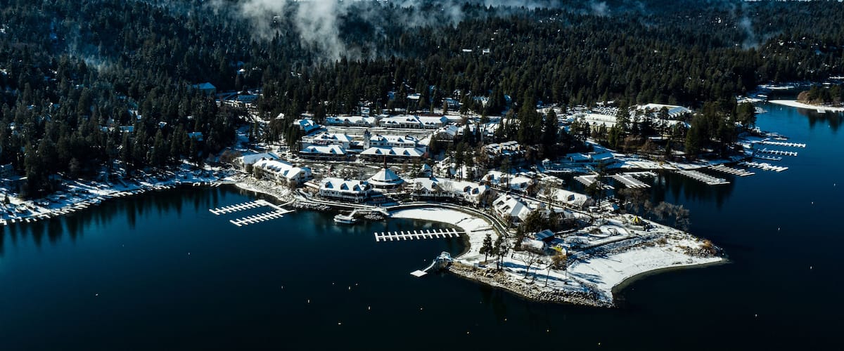An aerial view of a winter resort with a lake, snow-covered docks, and forested hills.
