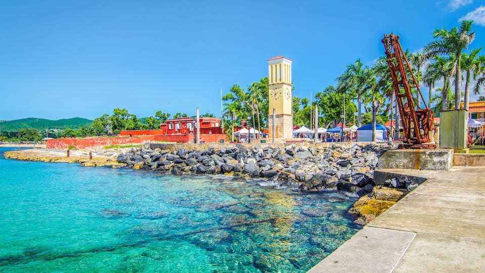A coastal town with a clear blue sea, a stone pier, a clock tower, and palm trees.
