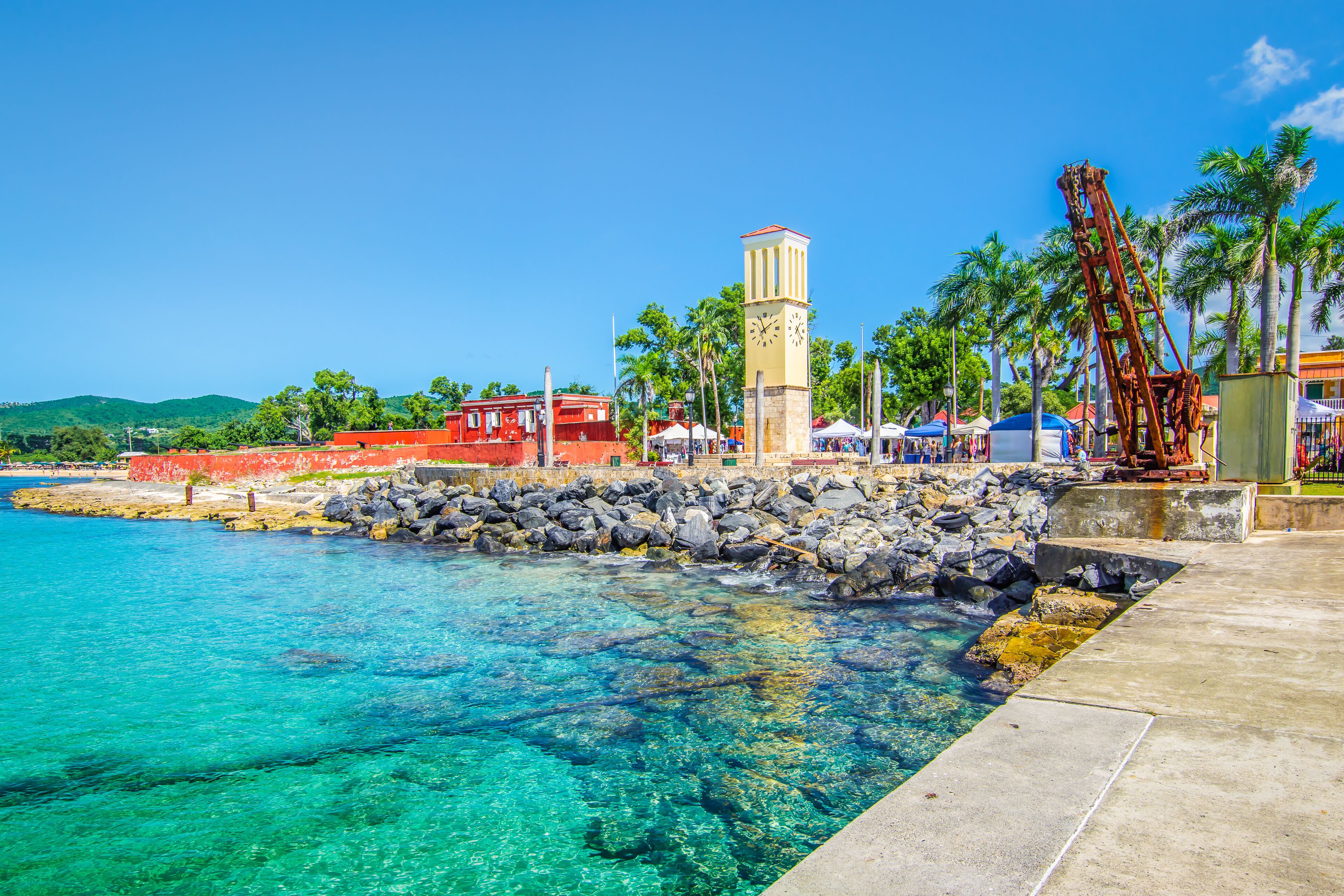 A coastal town with a clear blue sea, a stone pier, a clock tower, and palm trees.
