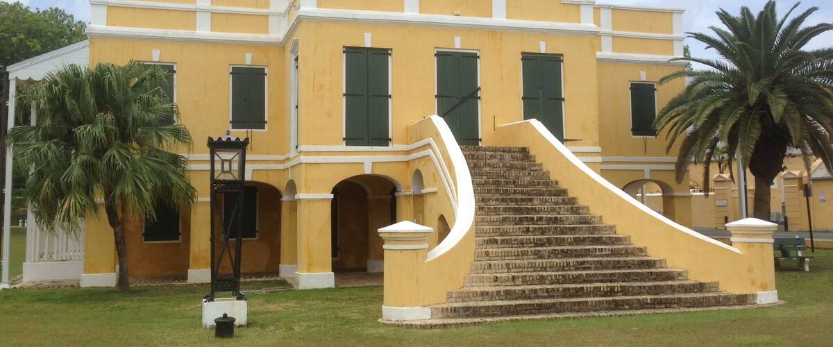 A yellow building with green shutters, a palm tree, and a staircase leading to the entrance.