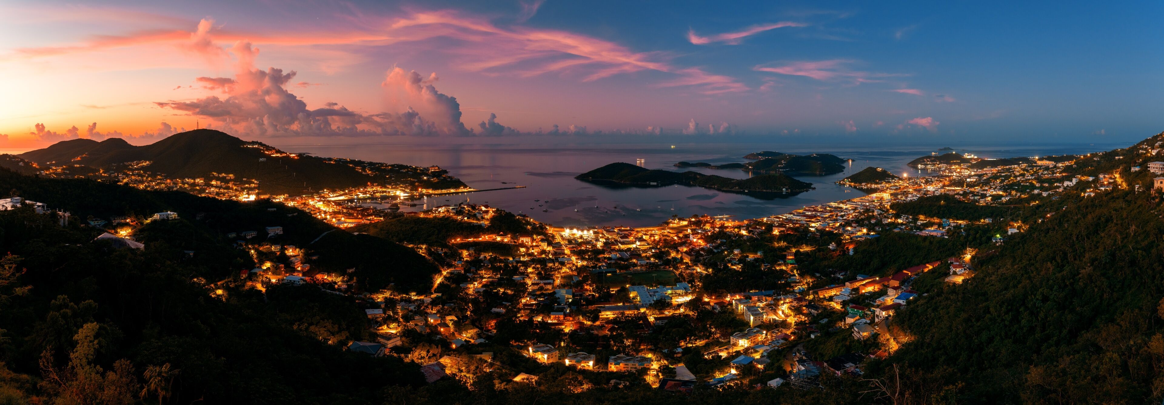 A coastal town at dusk with illuminated buildings, a bridge, and distant mountains.