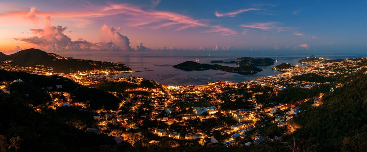 A coastal town at dusk with illuminated buildings, a bridge, and distant mountains.