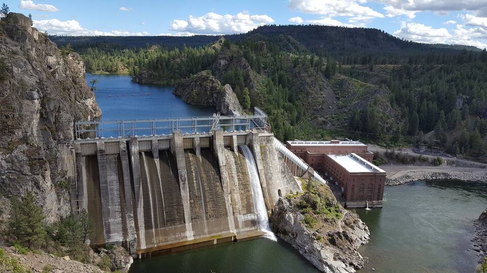 A dam with water flowing over its spillway, surrounded by forested hills and a clear sky.
