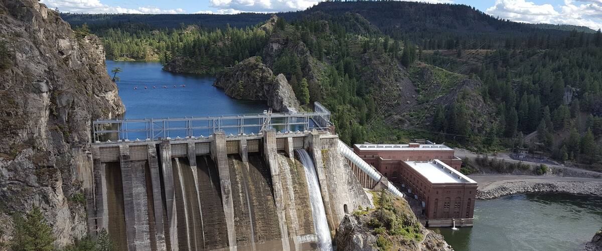 A dam with water flowing over its spillway, surrounded by forested hills and a clear sky.