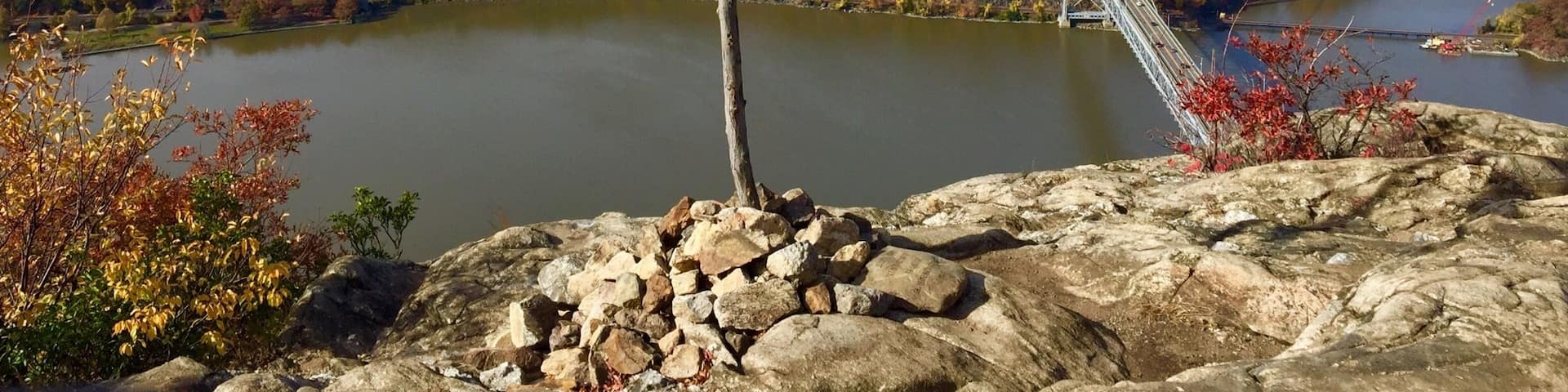 An American flag on a rocky outcrop overlooking a river, with a bridge and autumn foliage in the background.
