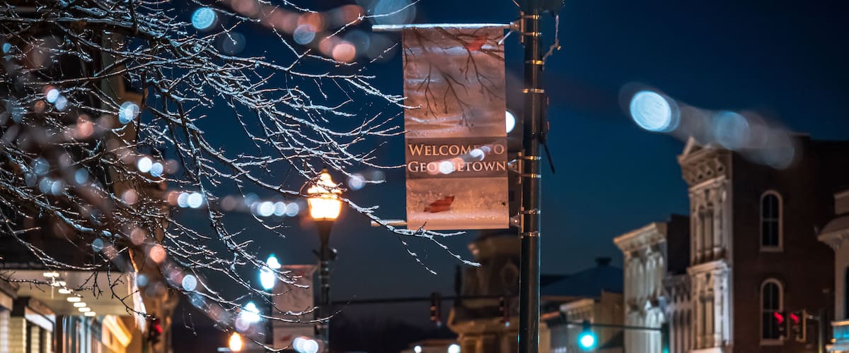 A snow-covered street with a 'Welcome to Georgetown' sign, illuminated street lamps, and historic buildings.