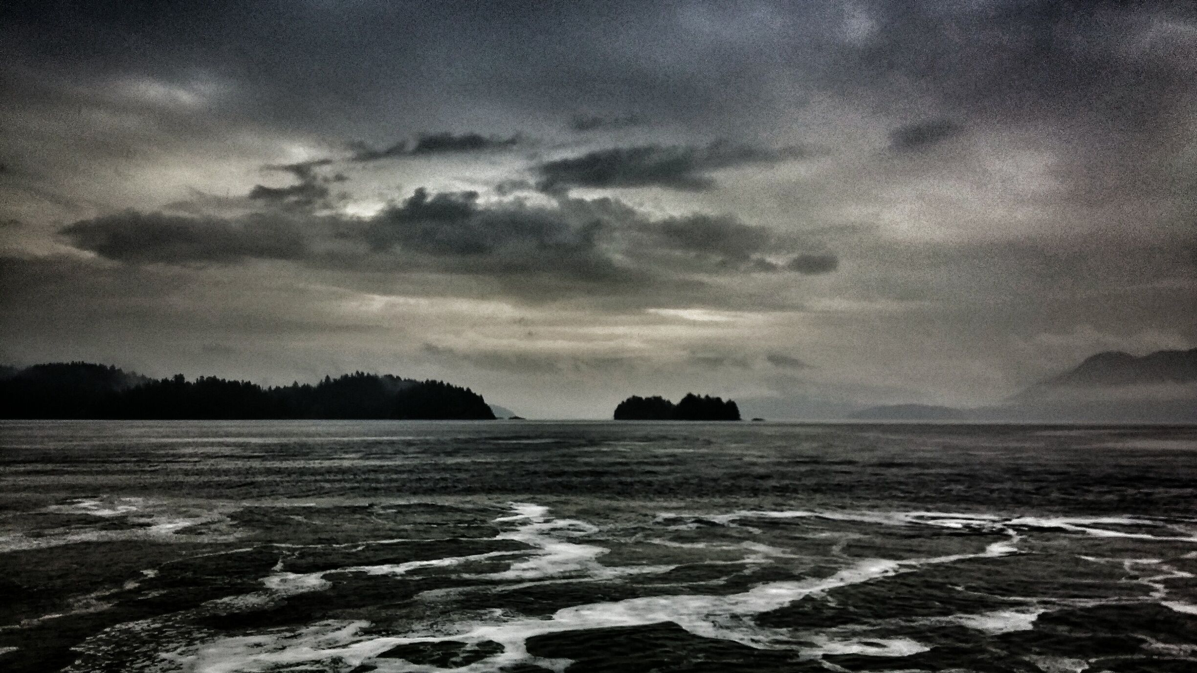 A stormy sea with waves crashing onto a rocky shore and distant islands under a cloudy sky.
