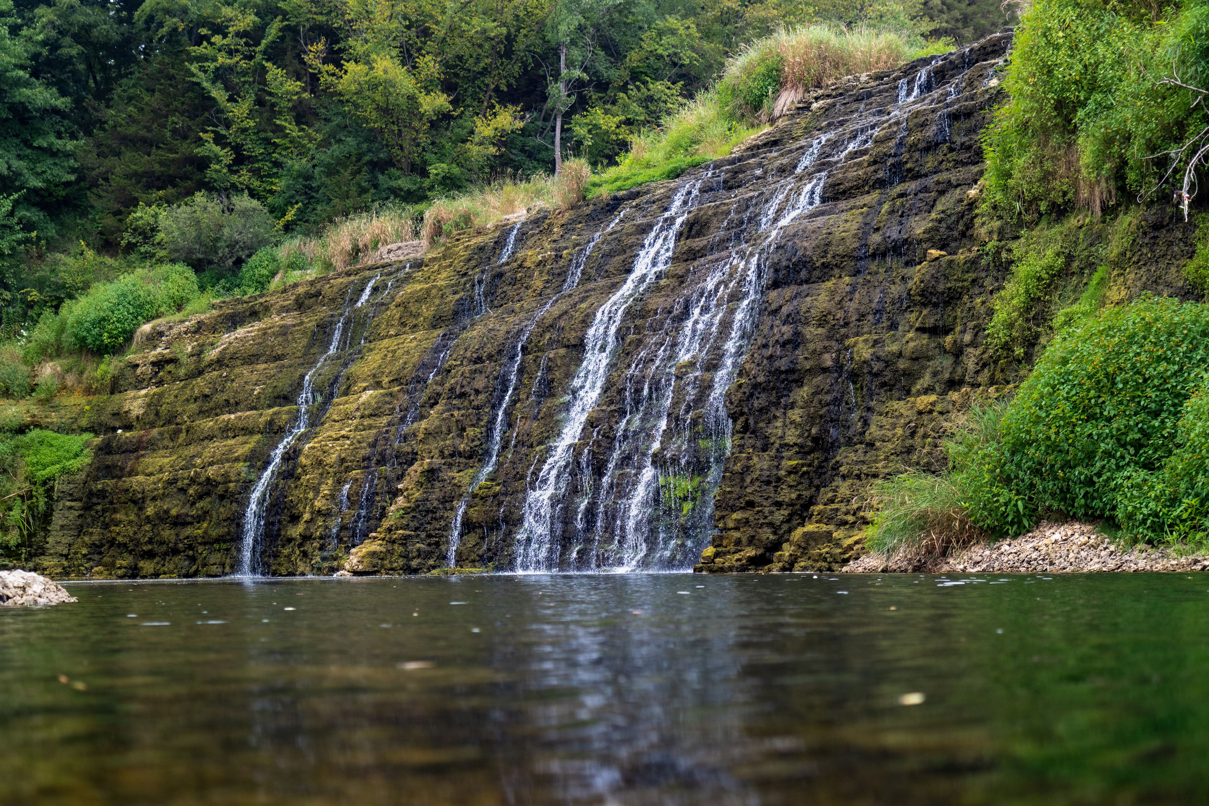 A multi-tiered waterfall cascading into a calm body of water, surrounded by lush greenery and dense forest.