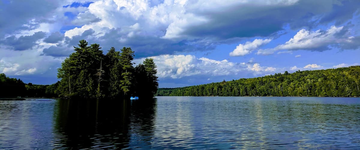 A calm lake with a small island of trees in the center, surrounded by a forested shoreline under a partly cloudy sky.