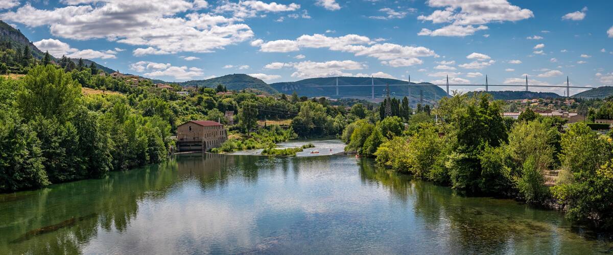 A river with a bridge in the background, surrounded by greenery and hills.