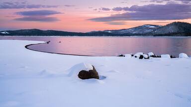 A snow-covered landscape with a frozen lake, a rock in the foreground, and a forested hill in the background.