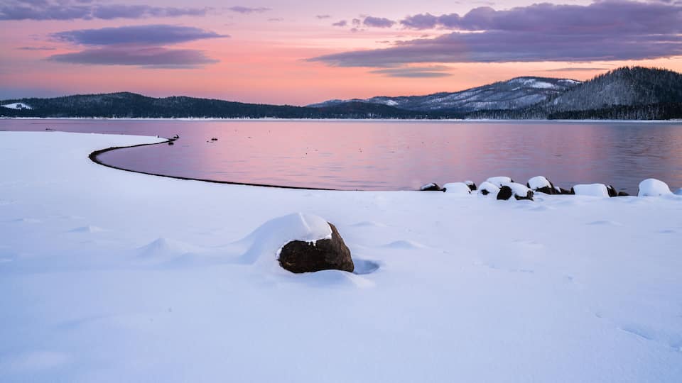 A snow-covered landscape with a frozen lake, a rock in the foreground, and a forested hill in the background.