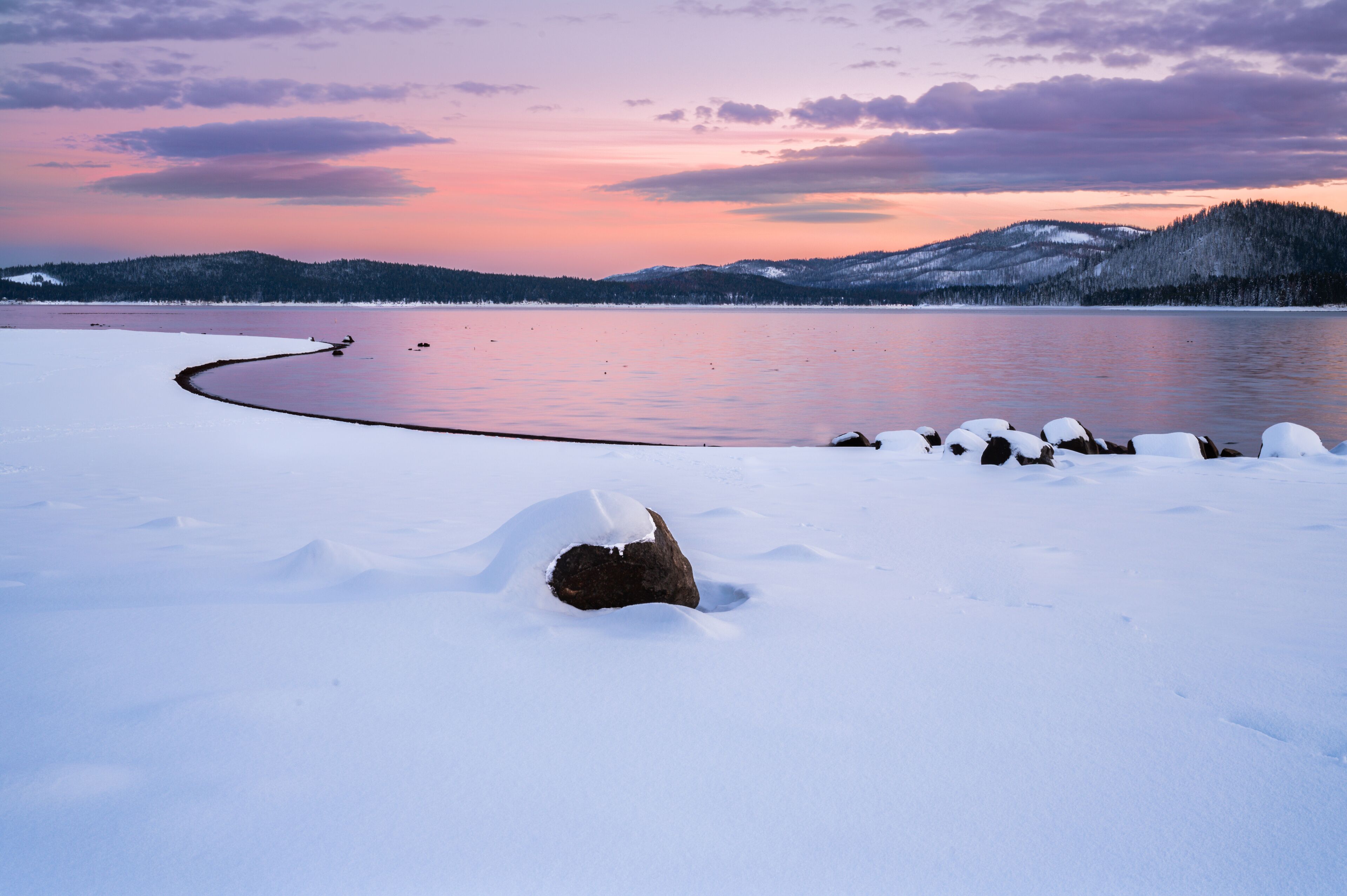 A snow-covered landscape with a frozen lake, a rock in the foreground, and a forested hill in the background.