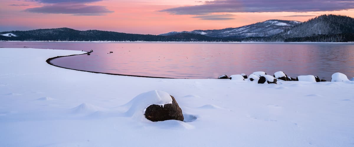 A snow-covered landscape with a frozen lake, a rock in the foreground, and a forested hill in the background.