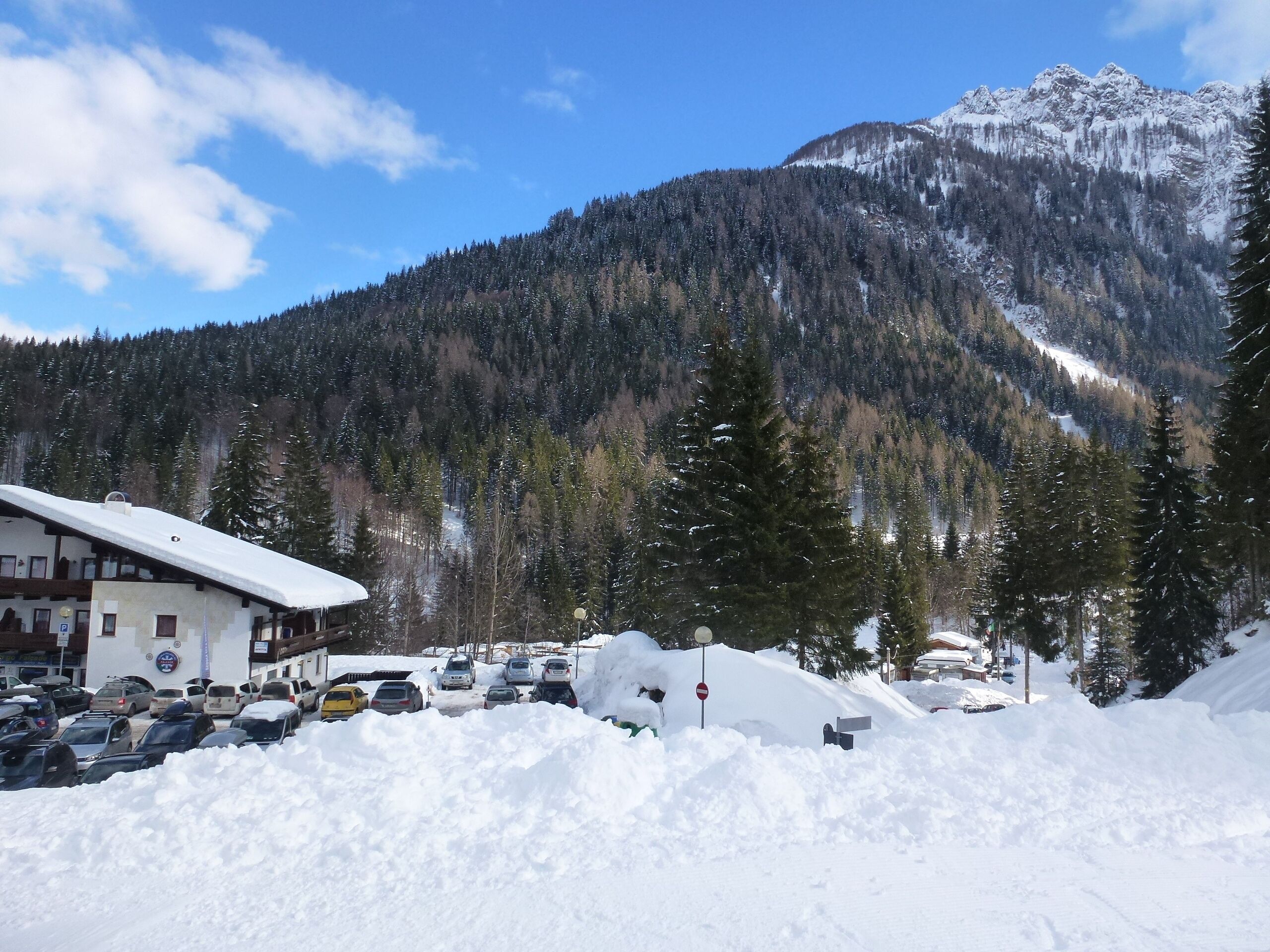 A snow-covered parking lot with cars and a building in the foreground, surrounded by snow-laden trees and a mountain in the background.