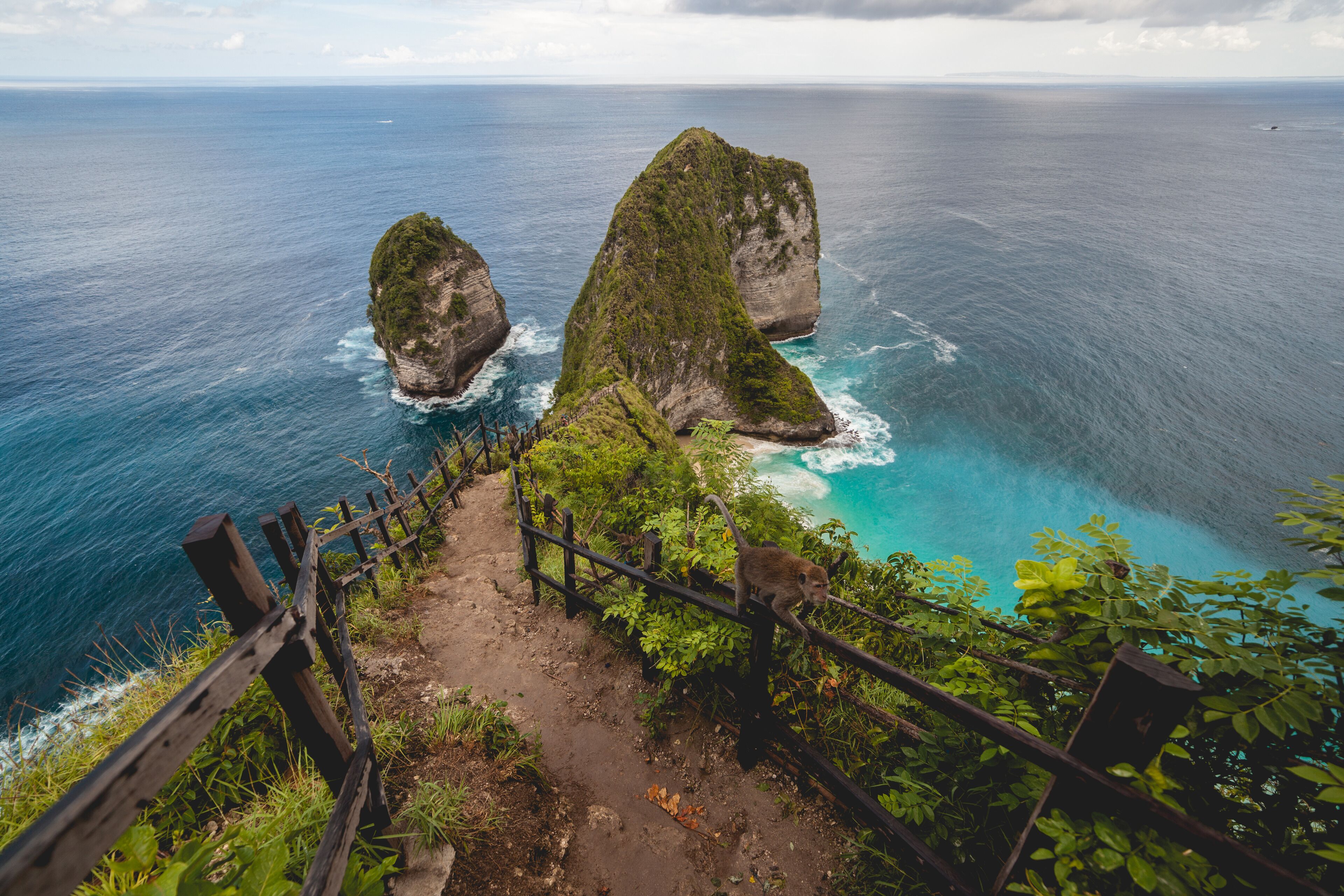 A coastal landscape with a wooden staircase leading down to the sea, surrounded by lush greenery and two prominent rock formations.