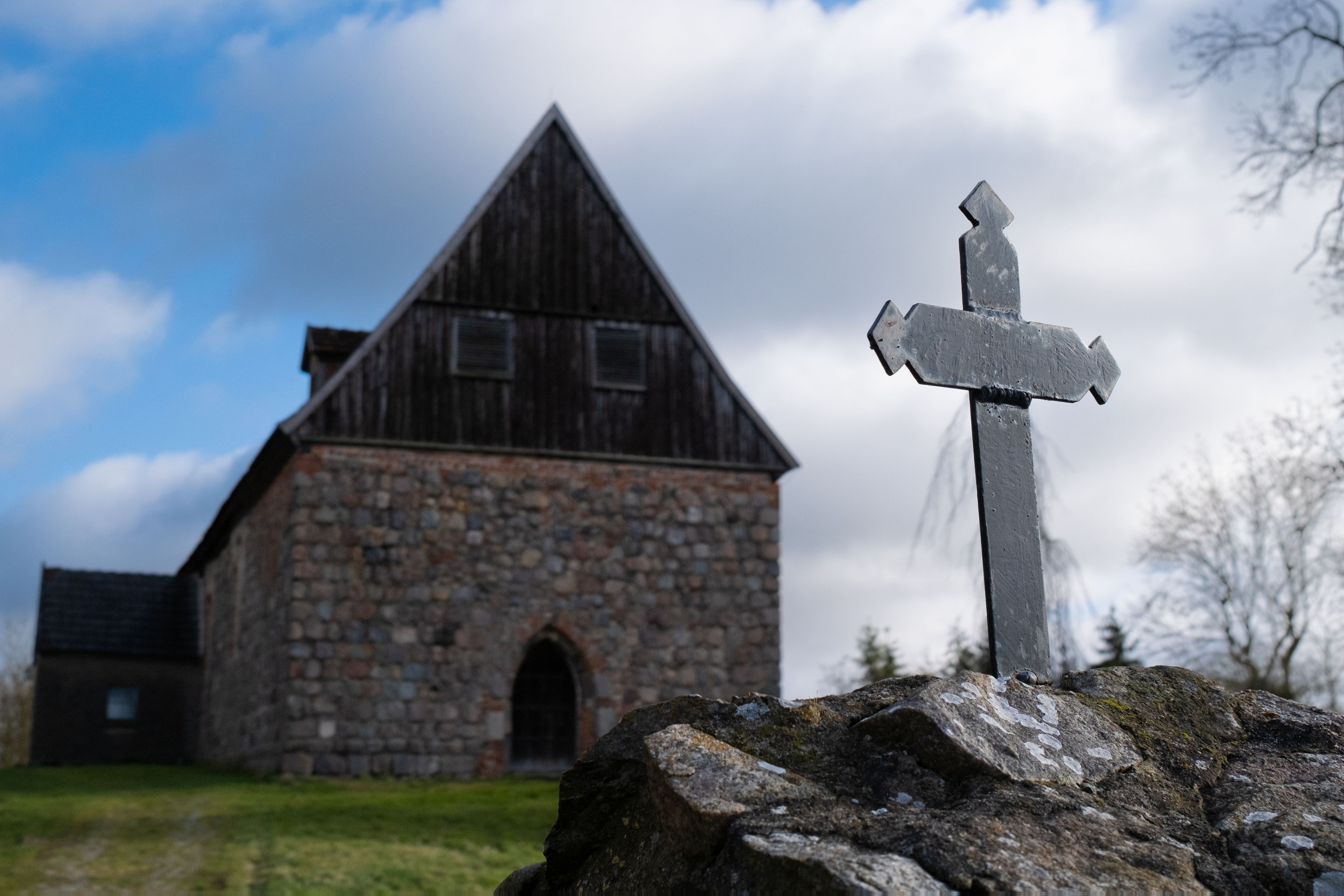 A stone church with a wooden steeple and a weathered cross in the foreground.