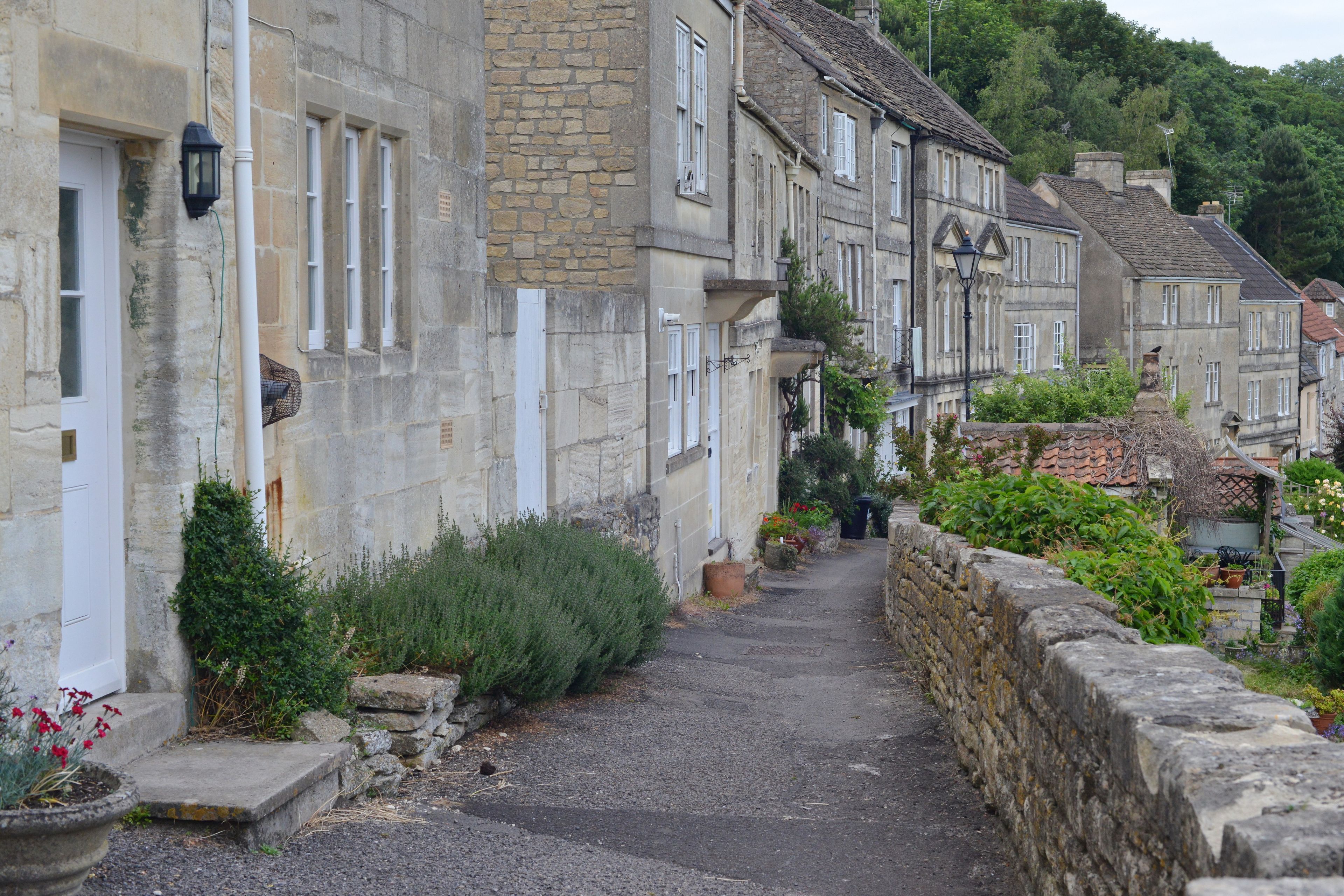A narrow cobblestone street flanked by historic stone buildings with white window frames and shutters, leading up a gentle slope.