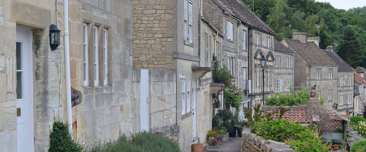 A narrow cobblestone street flanked by historic stone buildings with white window frames and shutters, leading up a gentle slope.