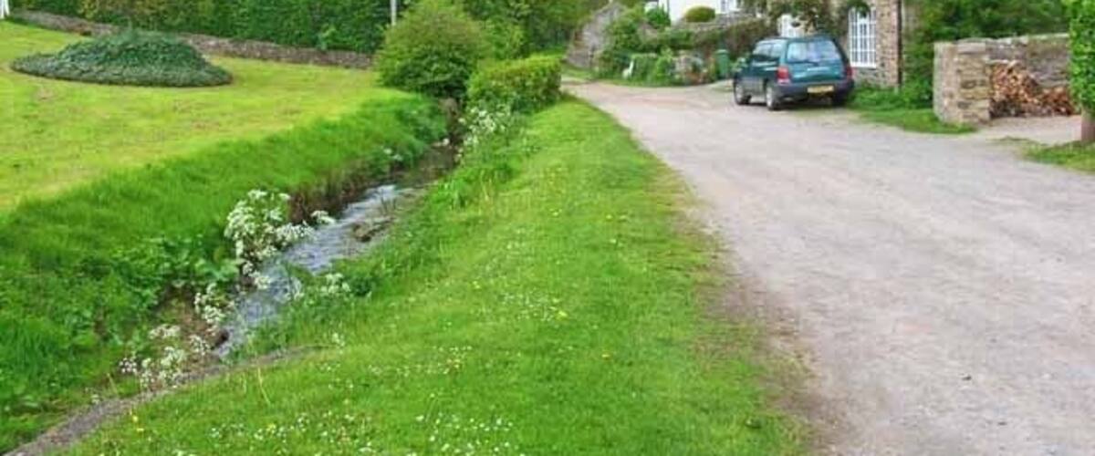 A rural road with a car parked on the side, surrounded by greenery and stone houses.