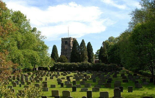 A cemetery with numerous headstones, surrounded by lush greenery and a prominent tower structure in the background.