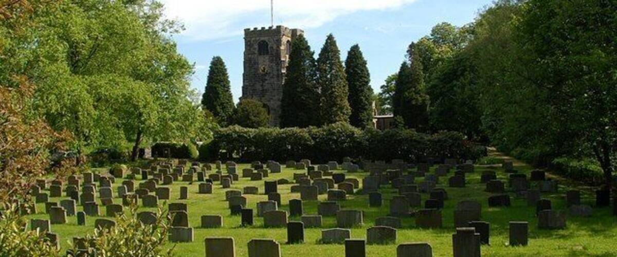 A cemetery with numerous headstones, surrounded by lush greenery and a prominent tower structure in the background.