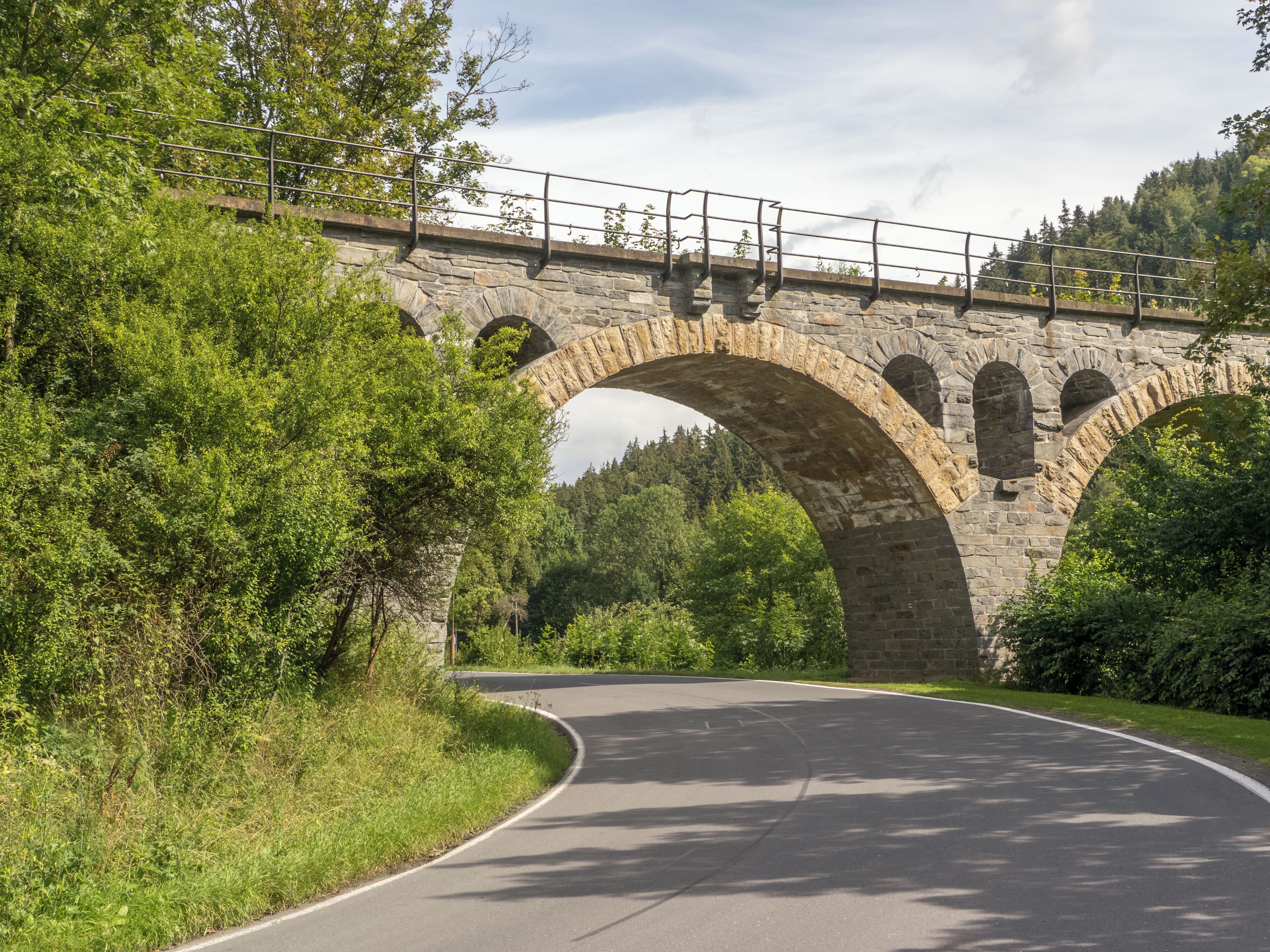 A stone bridge with multiple arches over a curved road, surrounded by lush greenery.