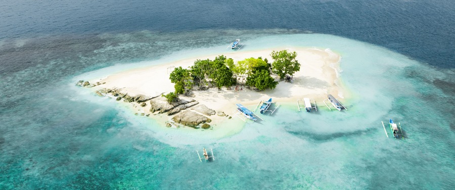An aerial view of a tropical island with clear turquoise waters, a sandy beach, lush green trees, and several boats anchored near the shore.
