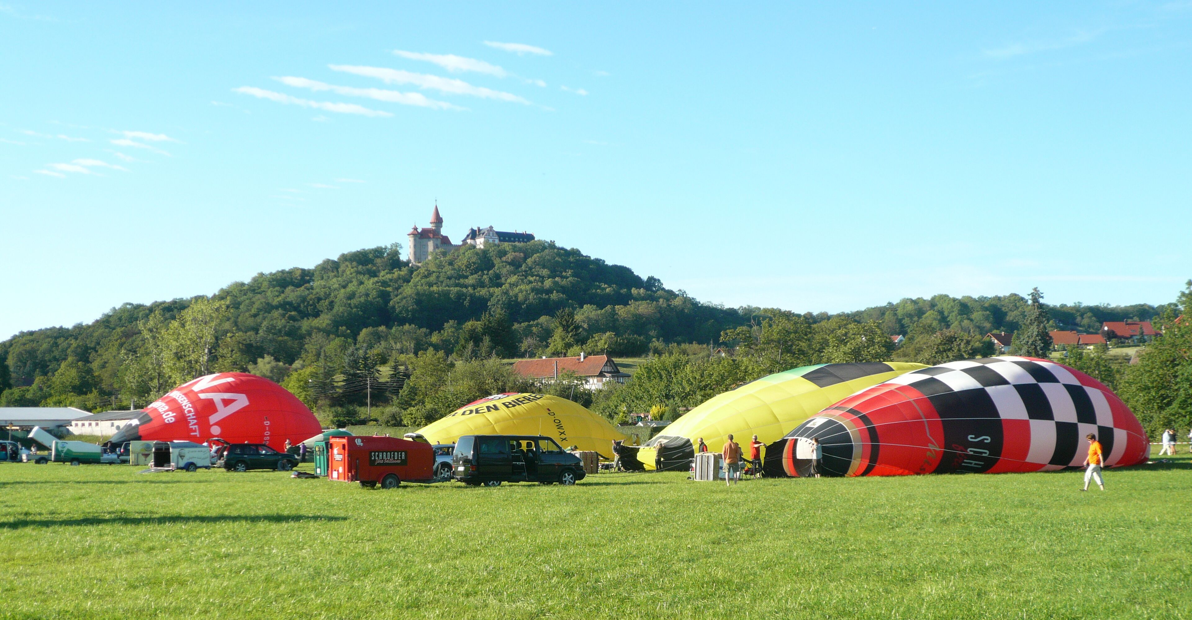 A field with several large hot air balloons, a castle on a hill in the background, and a clear blue sky.