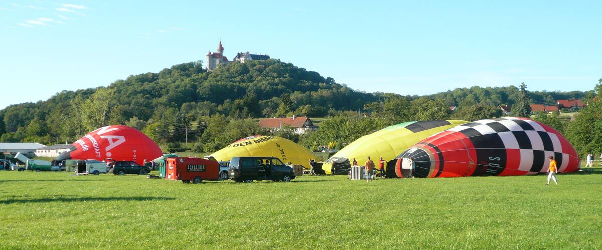 A field with several large hot air balloons, a castle on a hill in the background, and a clear blue sky.