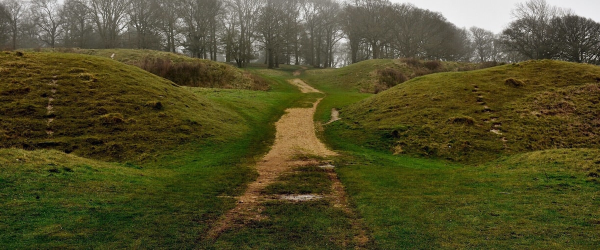 A narrow dirt path leading through a grassy field flanked by mounds of earth, with leafless trees in the background.
