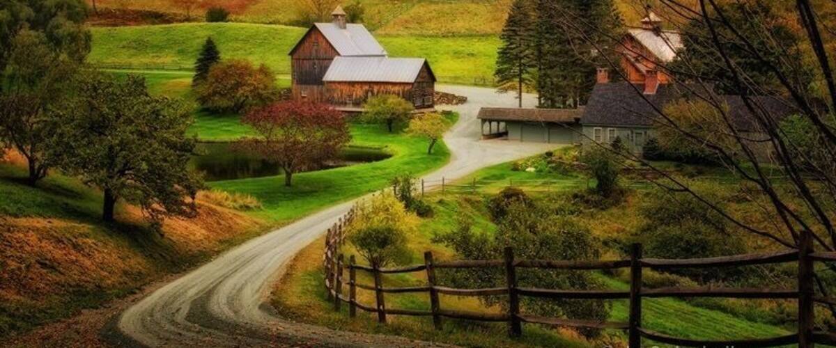 A rural landscape with a dirt road, wooden fence, barns, and a pond.