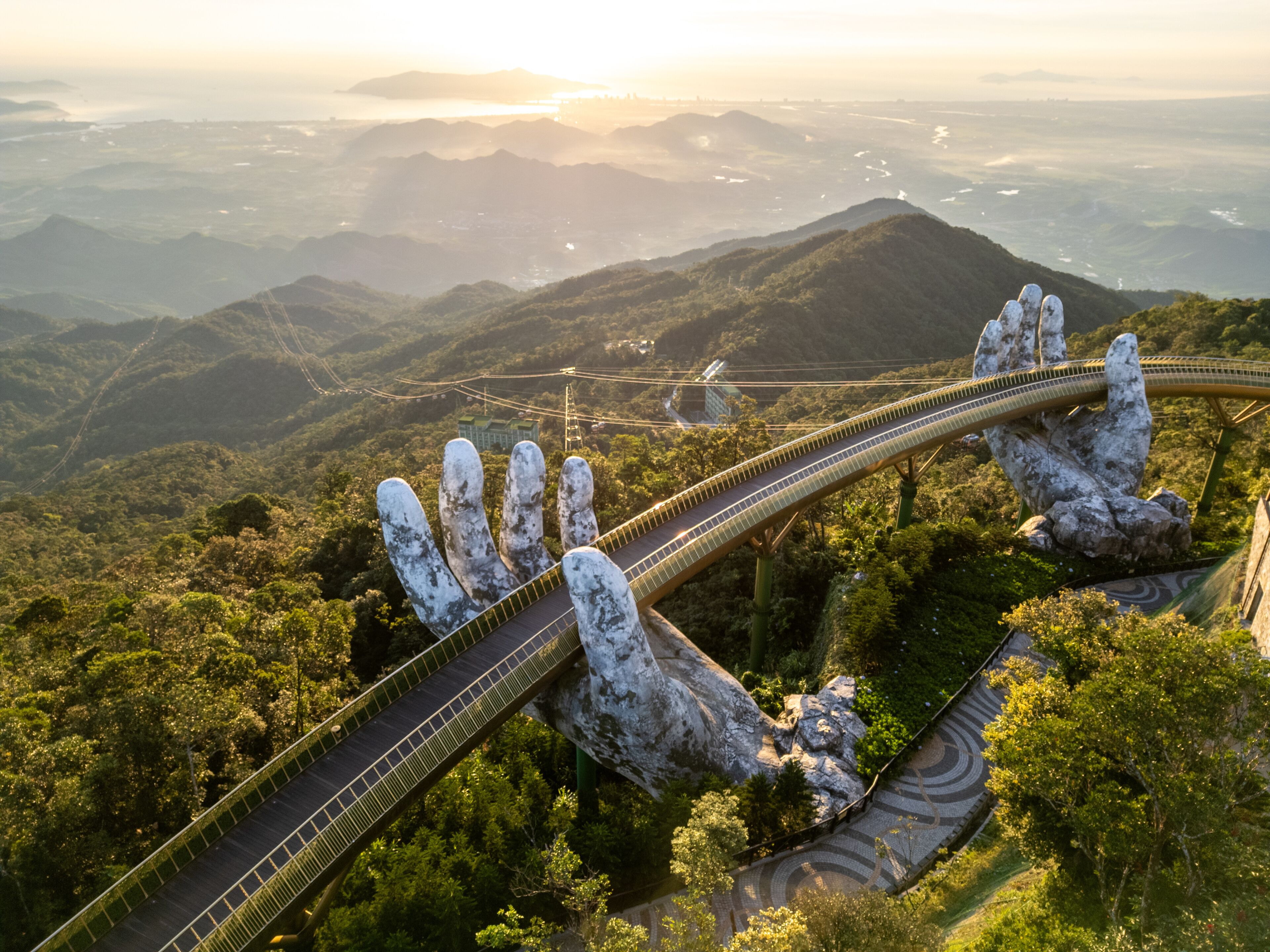 Aerial view of the iconic Golden Bridge, held by giant stone hands, stretching across the lush, sun-kissed mountains, DaNang, Da Nang, Vietnam.