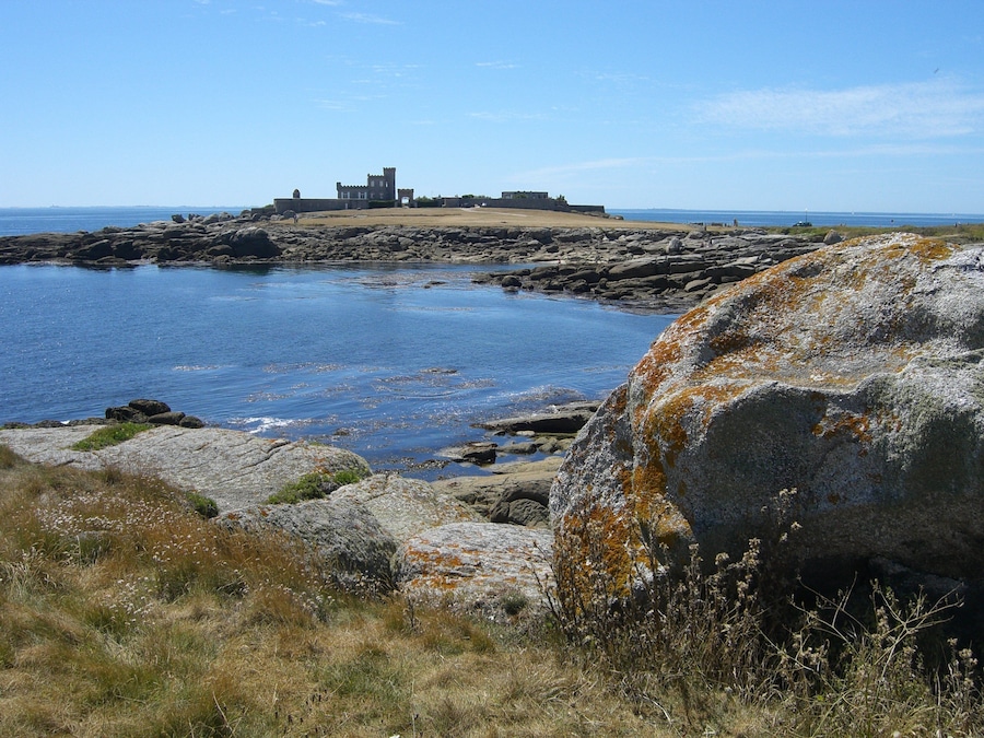 A coastal landscape with a historic stone building on a rocky outcrop, surrounded by calm waters and sparse vegetation.