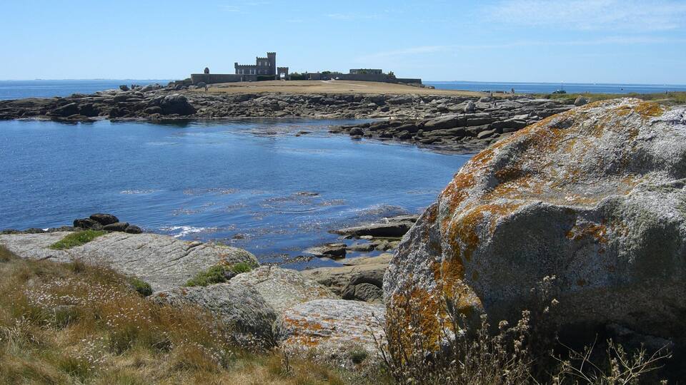 A coastal landscape with a historic stone building on a rocky outcrop, surrounded by calm waters and sparse vegetation.