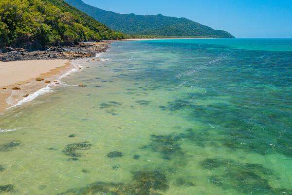 A beach with clear turquoise water, sandy shore, and a forested hill in the background.