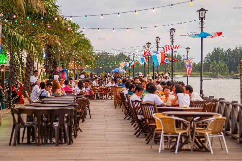 A crowded riverside dining area with tables and chairs, string lights, and flags.