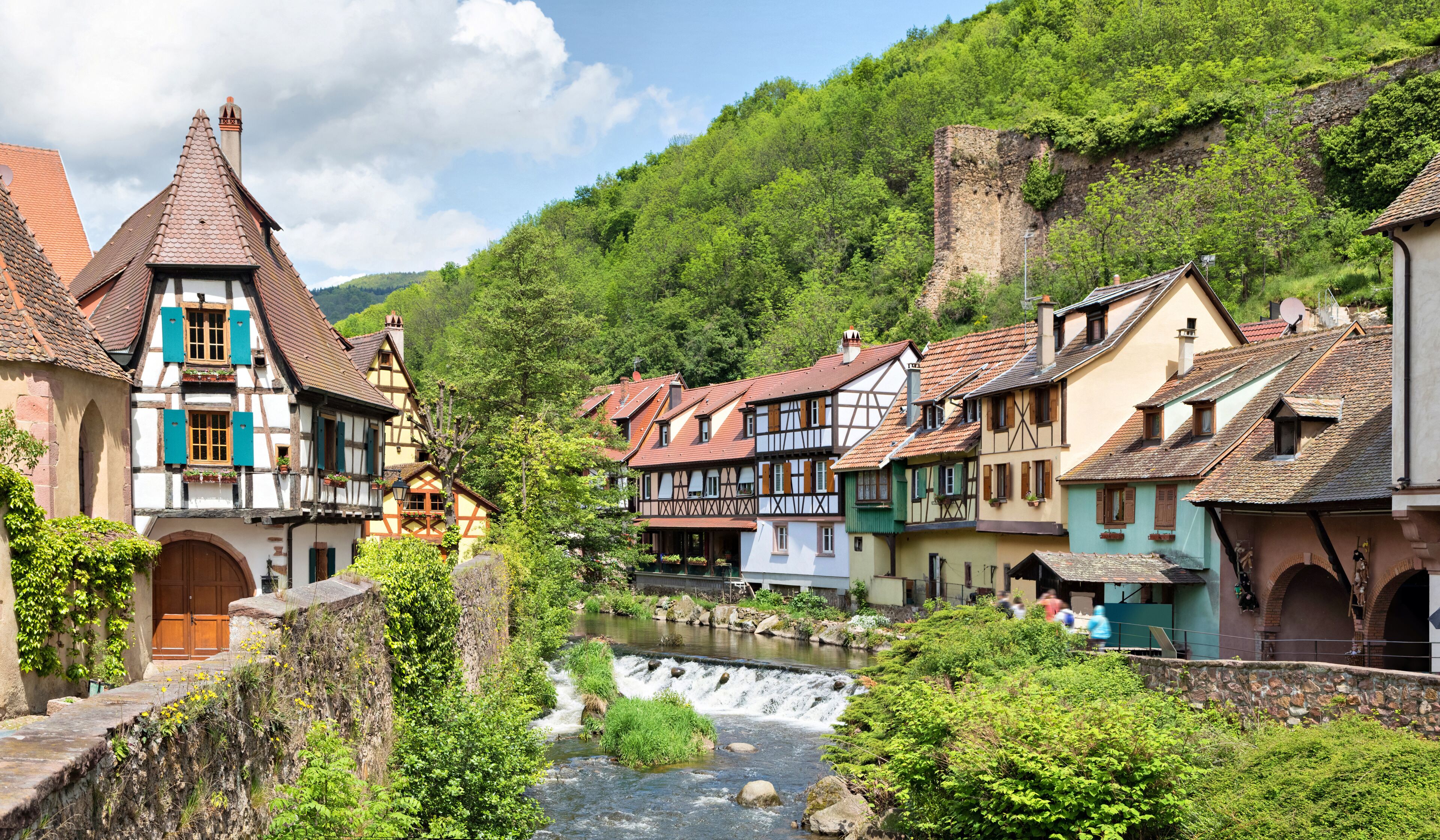 A picturesque European village with traditional half-timbered houses, a flowing river, and a castle on a hill.