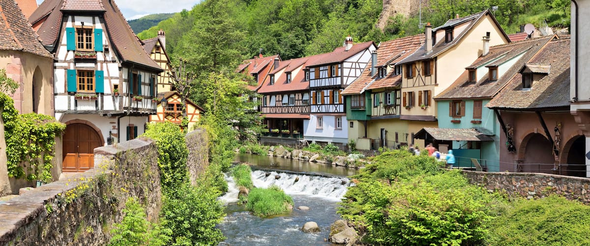 A picturesque European village with traditional half-timbered houses, a flowing river, and a castle on a hill.